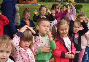 Przedszkolaki stoją w ogrodzie przedszkolnym. Niektóre z nich podnoszą ręce.