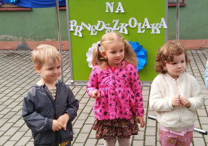 Dziewczynki i chłopiec stoją na tarasie przedszkolnym na tle dekoracji Pasowania na Przedszkolaka.