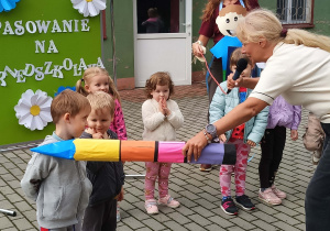 Pani Dyrektor pasuje na przedszkolaka dzieci dużą kredką. Nauczycielka przytrzymuje kabel od mikrofon.