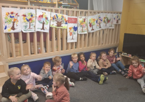 Przedszkolaki siedzą na dywanie. Nad nimi wiszą prace plastyczne - kolorowe drzewa.
