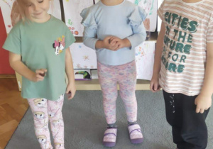 Troje dzieci stoi na dywanie trzymając w dłoniach kasztany. Przed nimi stoi koszyk z kasztanami.