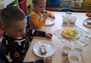 Chłopiec i dziewczynka siedzą przy stole, jedzą ciasto dyniowe. Na środku stołu stoją pokrojone jabłka i winogrono.