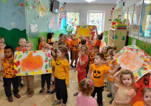 Przedszkolaki ubrane na pomarańczowo prezentują prace plastyczne - dynię w ramce z papierowych liści.
