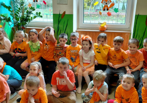 Przedszkolaki ubrane na pomarańczowo siedzą na ławce i na podłodze.
