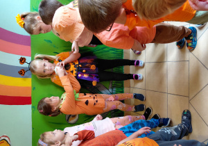 Przedszkolaki ubrane na pomarańczowo podają sobie dynię z gliny.