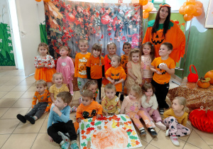 Przedszkolaki z grupy I ubrane na pomarańczowo wraz z nauczycielką pozują do zdjęcia na jesiennym tle.