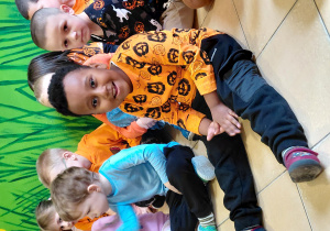 Chłopiec w pomarańczowej koszulce siedzi na podłodze. W tle inne dzieci.