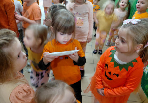 Dziewczynka wylizuje miód z talerza. Obok stoi dziewczynka ubrana na pomarańczowo. W tle dzieci.