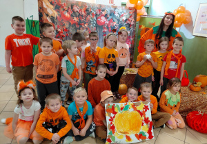 Przedszkolaki z grupy II ubrane na pomarańczowo wraz z nauczycielką pozują do zdjęcia na jesiennym tle.