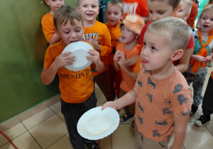 Dwóch chłopcow ubranych na pomarańczowo wylizuje miód z talerzy. W tle inne przedszkolaki ubrane na pomarańczowo.
