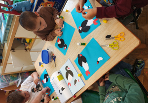 Chłopcy układają pingwiny z kół origami na niebieskich kartkach. Na stole leży książka z instrukcją wykonania pingwinów z kół origami.