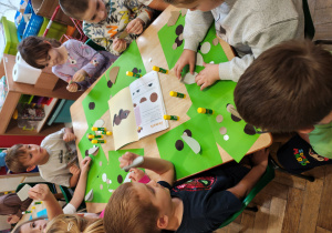 Przedszkolaki siedzą przy stole i wykonują jamnika z kół origami. Na stole leży książka z instrukcją wykonania jamnika z kół origami.