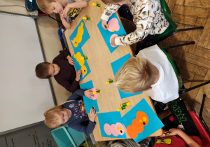 Przedszkolaki siedzą przy stole. Przed nimi leżą niebieskie kartki, koła origami i kleje.