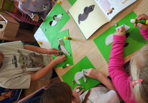 Przedszkolaki siedzą przy stole i wykonują jamnika z kół origami. Na stole leży książka z instrukcją wykonania jamnika z kół origami.