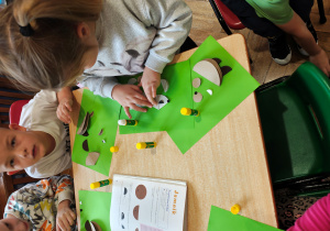 Przedszkolaki siedzą przy stole i wykonują jamnika z kół origami. Na stole leży książka z instrukcją wykonania jamnika z kół origami.