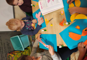 Przedszkolaki siedzą przy stole i wykonują ryby z kół origami. Na stole leży książka z instrukcją wykonania ryby z kół origami.