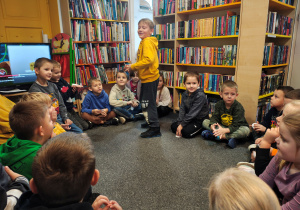 Przedszkolaki siedzą w kole na dywanie. Chłopiec stoi w środku koła.