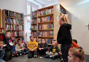 Przedszkolaki siedzą w kole na dywanie. Dziewczynka stoi w środku koła.