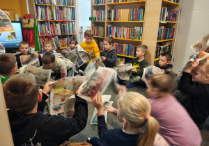 Przedszkolaki siedzą w kole na dywanie. W rękach trzymają rozłożone gazety.