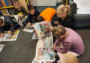 Przedszkolaki siedzą w kole na dywanie. Część dzieci w rękach trzymają rozłożone gazety, a część położyła gazety na dywanie.
