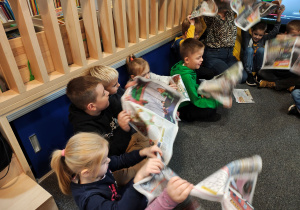 Przedszkolaki siedzą w kole na dywanie. W rękach trzymają rozłożone gazety.