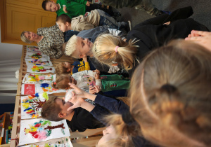 Przedszkolaki wraz z panią z biblioteki stoją w rozsypce na dywanie. Dmuchają w białe piórka.