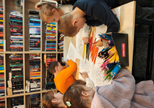 Przedszkolaki stoją przy stolikach i kolorują kredkami obrazek drzewa.