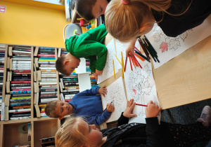 Przedszkolaki stoją przy stolikach i kolorują kredkami obrazek drzewa.