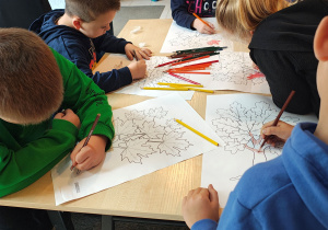 Przedszkolaki stoją przy stolikach i kolorują kredkami obrazek drzewa.