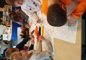 Przedszkolaki stoją przy stolikach i kolorują kredkami obrazek drzewa.