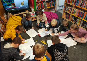 Przedszkolaki siedzą na dywanie i kolorują kredkami obrazek drzewa.