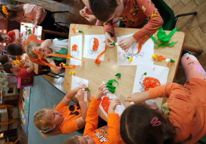 Przedszkolaki w pomarańczowych ubraniach siedzą przy stołach i wyklejają bibułą obrazki dyni.