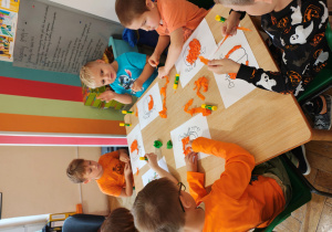 Przedszkolaki w pomarańczowych ubraniach siedzą przy stołach i wyklejają bibułą obrazki dyni.