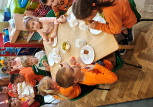 Przedszkolaki w pomarańczowych ubraniach siedzą przy stołach i jedzą ciasto dyniowe.