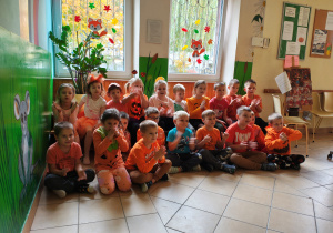 Przedszkolaki w pomarańczowych ubraniach siedzą na ławce i na podłodze w korytarzu przedszkola.