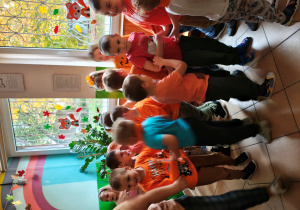 Przedszkolaki w pomarańczowych ubraniach stoją i tańczą w korytarzu przedszkola.