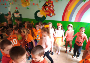 Przedszkolaki w pomarańczowych ubraniach stoją trzymając się za ręce i tańczą w korytarzu przedszkola.