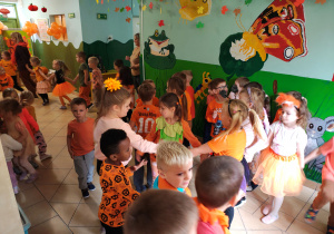 Przedszkolaki w pomarańczowych ubraniach stoją trzymając się za ręce i tańczą w korytarzu przedszkola.