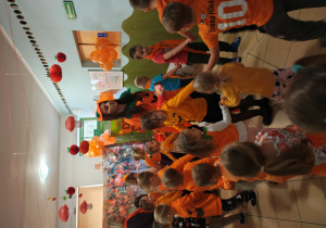 Przedszkolaki w pomarańczowych ubraniach stoją trzymając się za ręce i tańczą wraz z nauczycielką w korytarzu przedszkola.