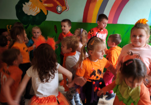 Przedszkolaki w pomarańczowych ubraniach stoją trzymając się za ręce i tańczą w korytarzu przedszkola.