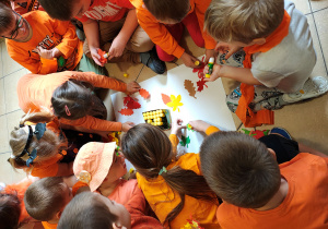 Przedszkolaki siedzą na podłodze i przyklejają do kartonu kolorowe liście z papieru.