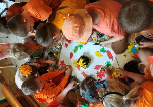 Przedszkolaki siedzą na podłodze i przyklejają do kartonu kolorowe liście z papieru.