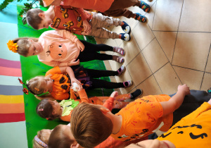 Przedszkolaki w pomarańczowych ubraniach stoją w kole i podają sobie dynię z gipsu.