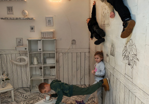 Chłopiec stoi przy stole z herbatą. Troje dzieci leży na ścianie, jeden chłopiec stoi na ścianie.