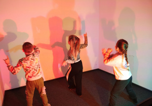 Dwie dziewczynki i chłopiec tańczą odwróceni tyłem. W tle kolorowe cienie dzieci.