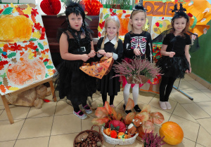 Dwie dziewczynki przebrane za Wednesday, dziewczynka przebrana za czarownicę i dziewczynka przebrana za nietoperza stoją trzymając jesienne dekoracje: dynię, wrzosy, halloweenowy koszyk. Przed i za dziewczynkami jesienna dekoracja.
