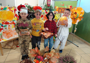 Chłopiec przebrany za rannego żołnierza, chłopiec przebrany za Jokera, chłopiec przebrany za Harrego Pottera i chłopiec przebrany za zawodnika judo stoją trzymając jesienne dekoracje: dynię, wrzosy, halloweenowy koszyk. Przed i za chłopcami jesienna dekoracja.