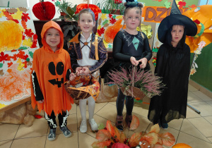 Chłopiec przebrany za dynię, dziewczynka przebrana za Rumi, dziewczynka przebrana za pająka i dziewczynka przebrana za czarownicę stoją trzymając jesienne dekoracje: dynię, wrzosy, halloweenowy koszyk. Przed i za dziećmi - jesienna dekoracja