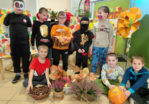 Trzech chłopców przebranych za zoombie, chłopiec za lego ninjago, chłopiec przebrany za dynię stoją, trzech chłopców bez przebrań siedzi trzymając jesienne dekoracje: dynię, wrzosy, halloweenowy koszyk. Przed i za chłopcami jesienna dekoracja.