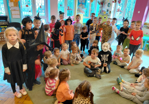 Przedszkolaki przebrane na Halloween oraz przedszkolaki bez przebrań stoją i siedzą na dywanie.